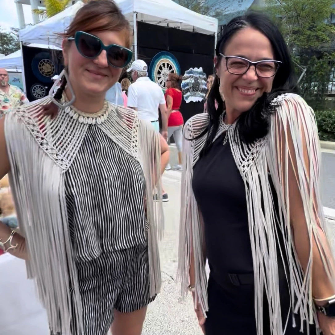 Two women wearing fringe outfits standing outdoors with a tent and people in the background.