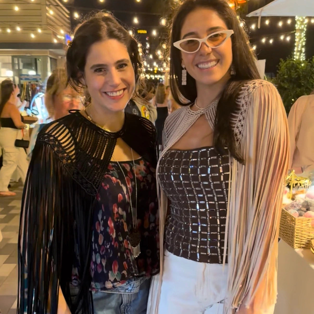 Two women posing together at an outdoor event with leia cape fringed top in beige and black