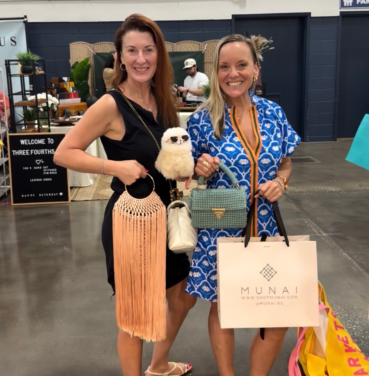 Two women smiling and posing together at an indoor event, each holding handbags. The woman on the left holds a peach macrame fringe bag. 