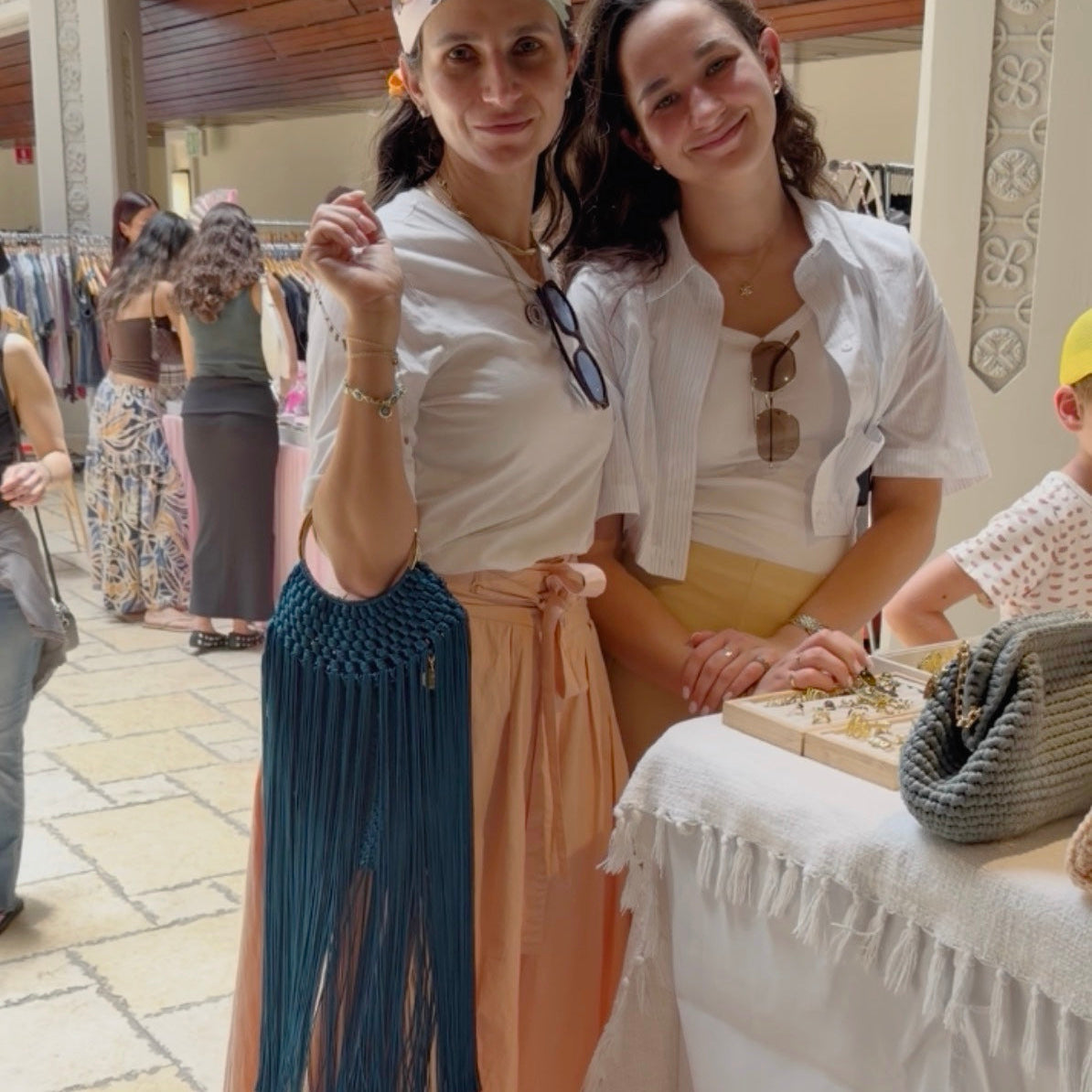 Two women posing together at what appears to be a boutique or market stall, showcasing bohemian-style clothing and accessories. The woman on the left wears a white t-shirt, a peach maxi skirt, a colorful headwrap, and holds a blue macrame fringe purse.