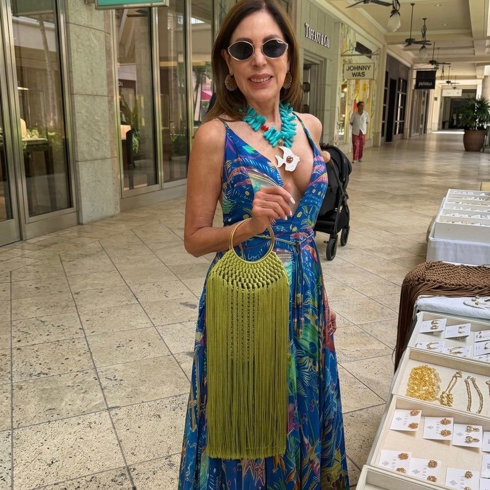 A woman wearing a vibrant blue tropical print maxi dress, turquoise necklace, and sunglasses stands in a shopping mall holding a lime green tiffany fringe handbag.