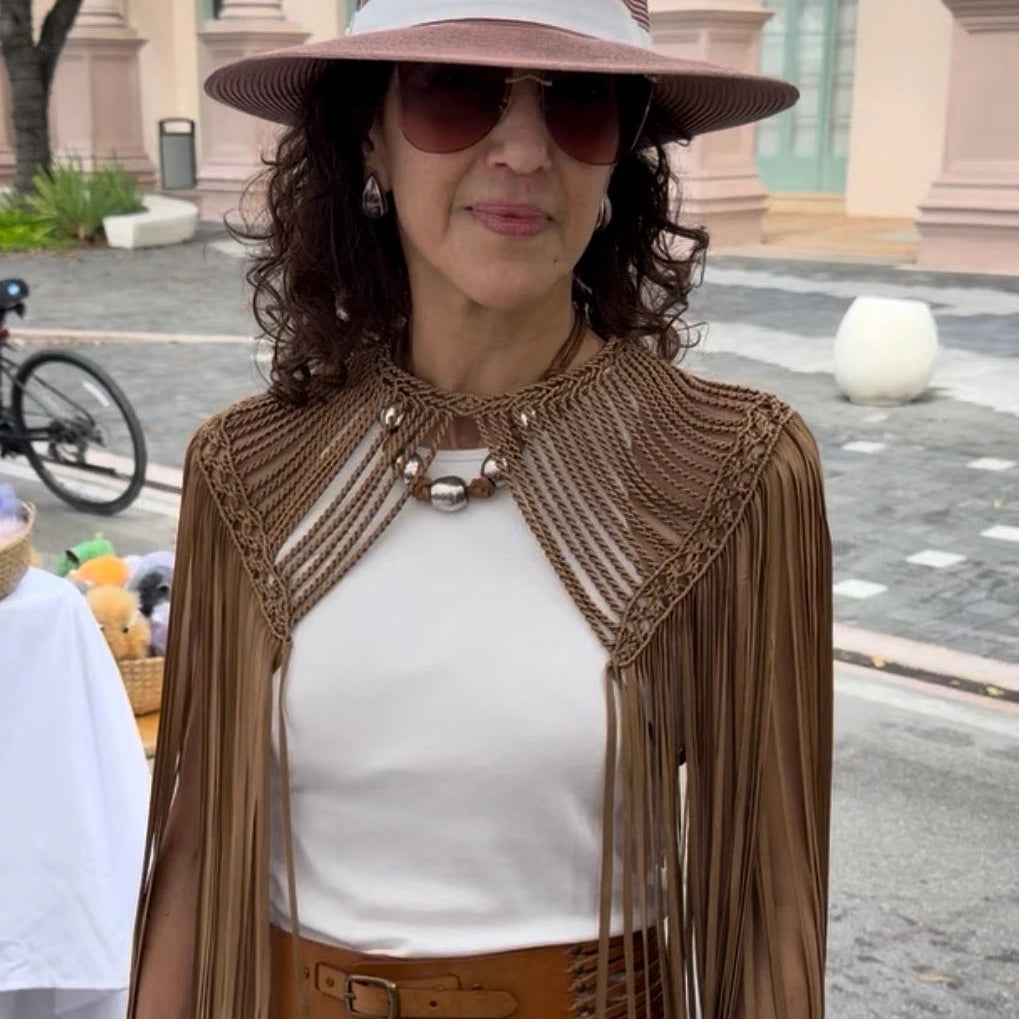 Woman wearing a brown fringed Leia Cape white top, and wide-brimmed hat outdoors. 
