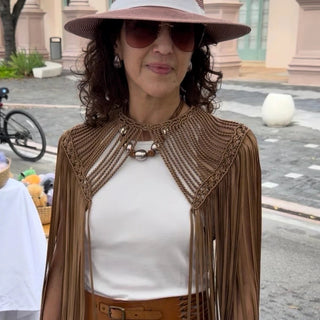 Woman wearing a brown fringed Leia Cape white top, and wide-brimmed hat outdoors. 